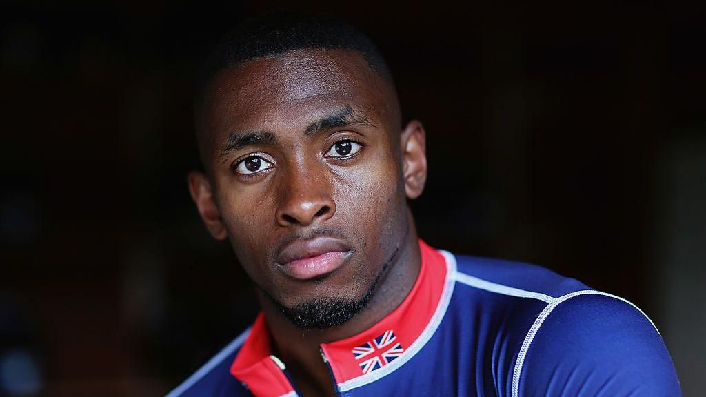 Joel Fearon is wearing a blue, red, and white jacket, with the Great Britain flag on the collar. He has black hair and black facial hair.