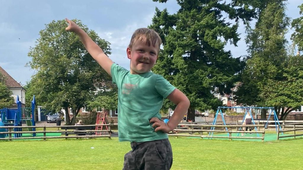 Toby standing on top of a box in a playground. He is posing and pointing one hand in the air. 