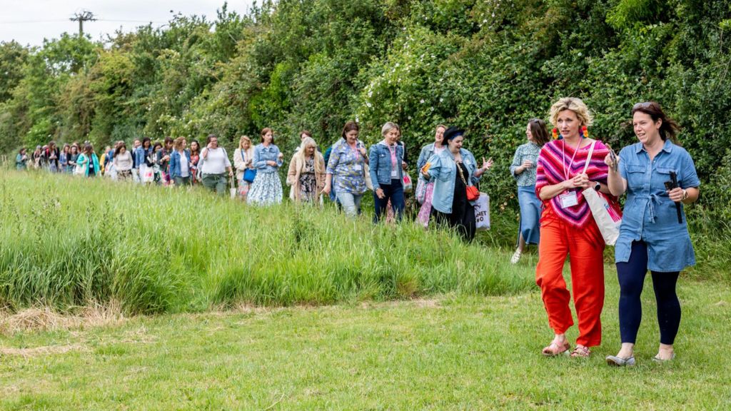 A long line of women walking in twos or threes, talking and smiling as they walk along a path near the edge of a field of long grass. It is taken at the Freelance Mum Mega Meet Up, which takes place at Tractors & Cream in Somerset.