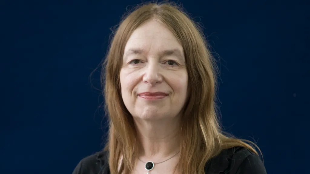 A woman with long brown hair and a black top poses in front of a dark blue background