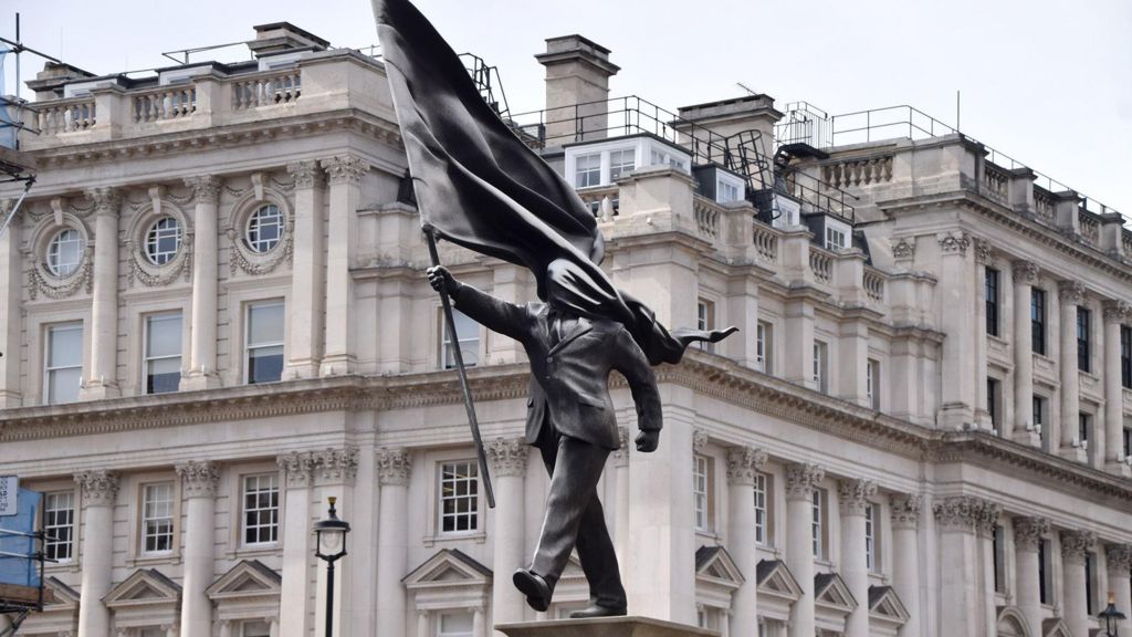 A grey statue of a man wearing a suit, walking forward, carrying a flag that covers his face