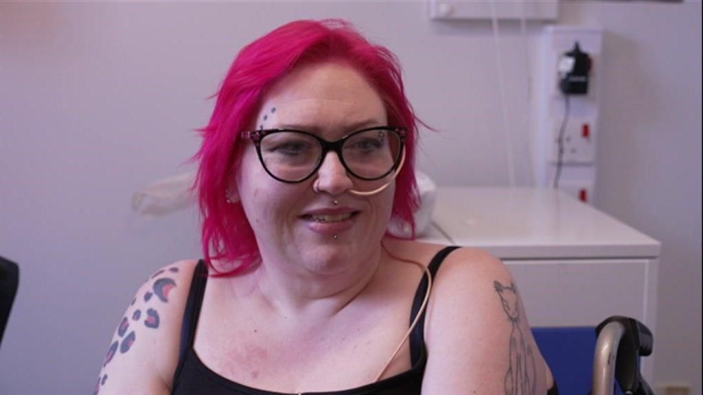 Pinky Jolley with bright pink hair, a black vest top, tattoos on her arms and a feeding tube in her nose