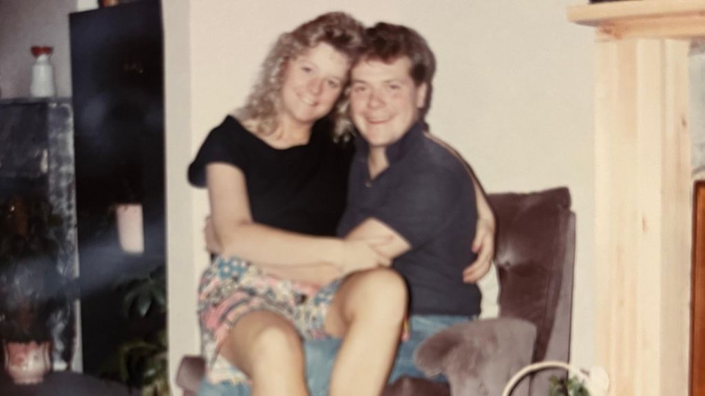 In an old picture, Sue sits on Alex's lap, both smiling. She has curly blonde hair down to her shoulders, wears a black T-shirt and colourful shorts. He sits in a soft brown/grey armchair and they have their arms around each other. He wears a grey/black T-shirt and jeans and they are in what looks like a living room. It is a slightly grainy picture of a picture. 