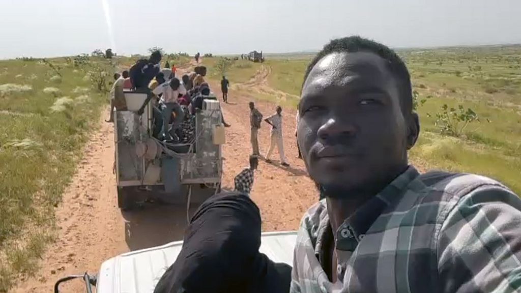 Mostafa, in a checked shirt, sits on top of a white vehicle which is driving along an untarmacked road. There is a van in front of his vehicle that is full of people and more people are walking along the road.