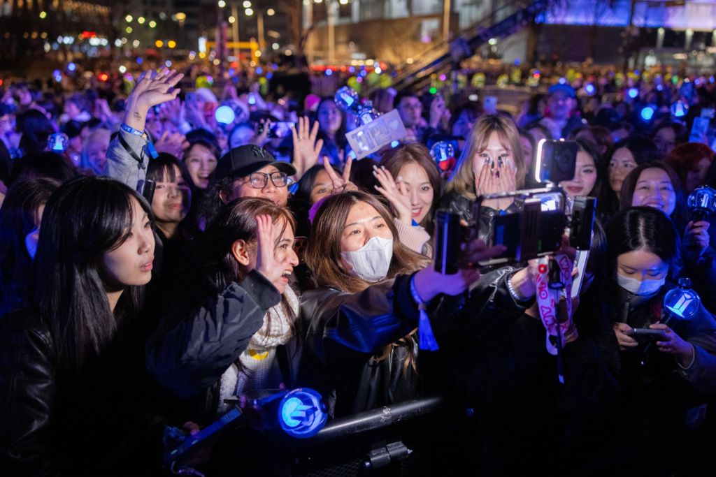 A dozen fans looking at a camera with a light above it, surrounded by purple light-sticks