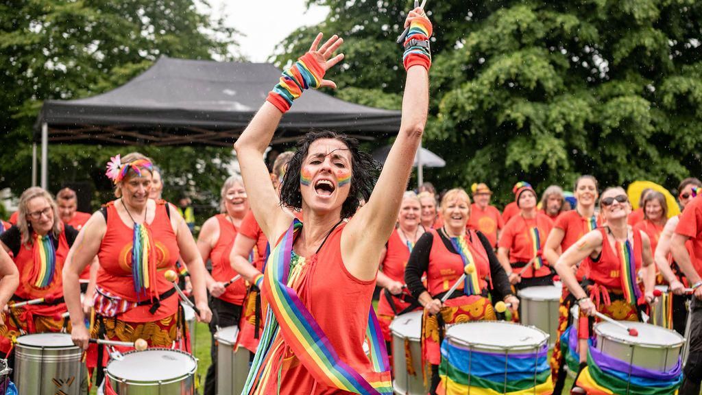 Women in red vests and rainbow sashes bang drums