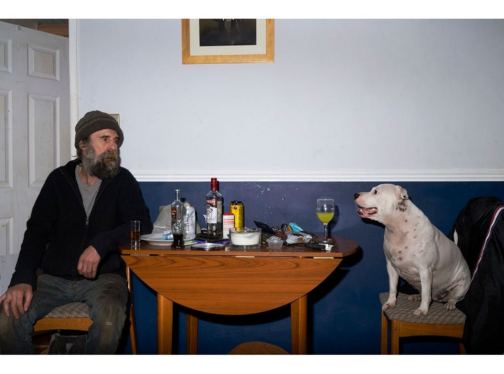 Man say on a chair with a table in between him and his dog, who is also sat on a chair. There are various beverages on the table