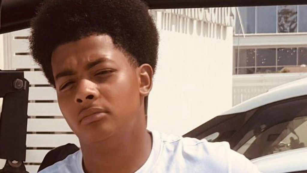 A teenage boy leaning against a car, wearing a white top
