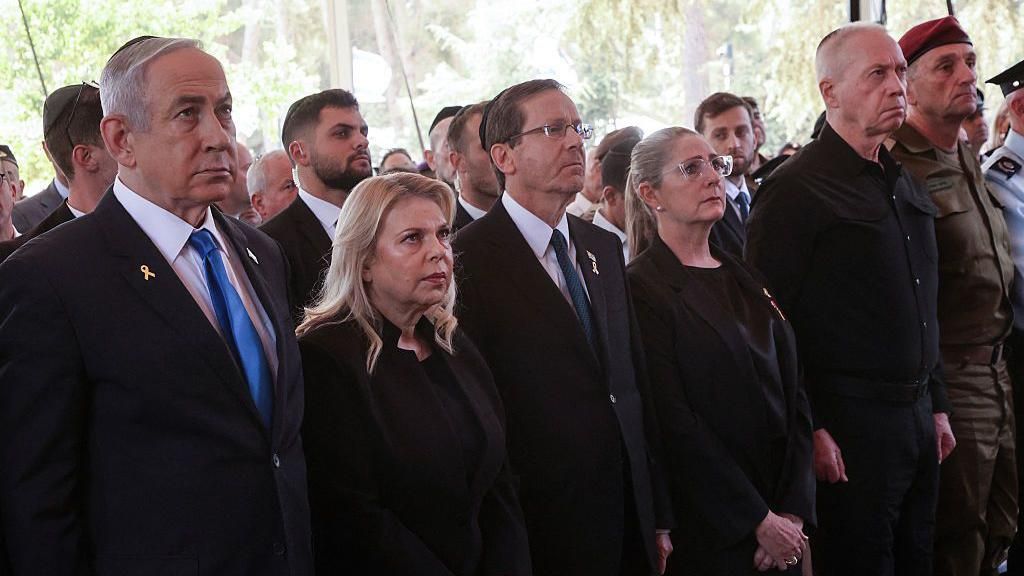 Netanyahu, Herzog and Gallant stand aside several others at a ceremony. The expressions are serious, or sombre, and most people are wearing suits or military attire. 