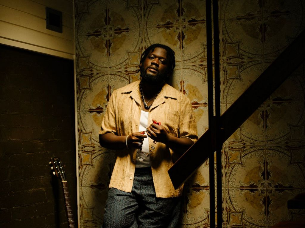 Michael Kiwanuka holds prayer beads, with a guitar propped up against the wall in a 1970s-themed house, in a promotional photo for his new music.