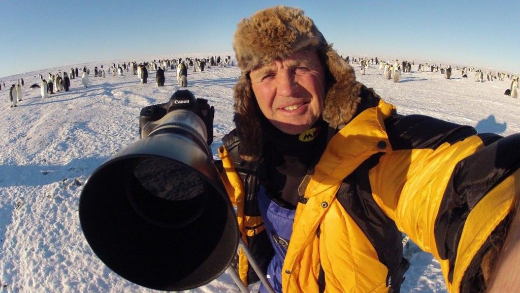 Doug Allan taking a selfie holding a large camera with many penguins on snow behind him