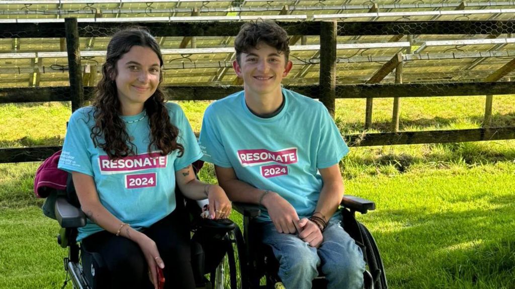 Molly and her brother Stan, in wheelchairs, with a background of a field and a lake. 