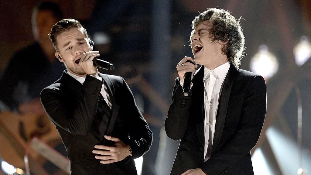 Liam Payne and Harry Styles performing together on stage. The pair both wear black and white suits and there is a band behind them