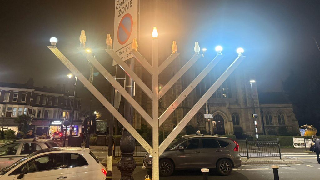 A giant Hanukkah menorah on display in front of a road junction, with some of the light bulbs representing candles on the top of the menorah illuminated.