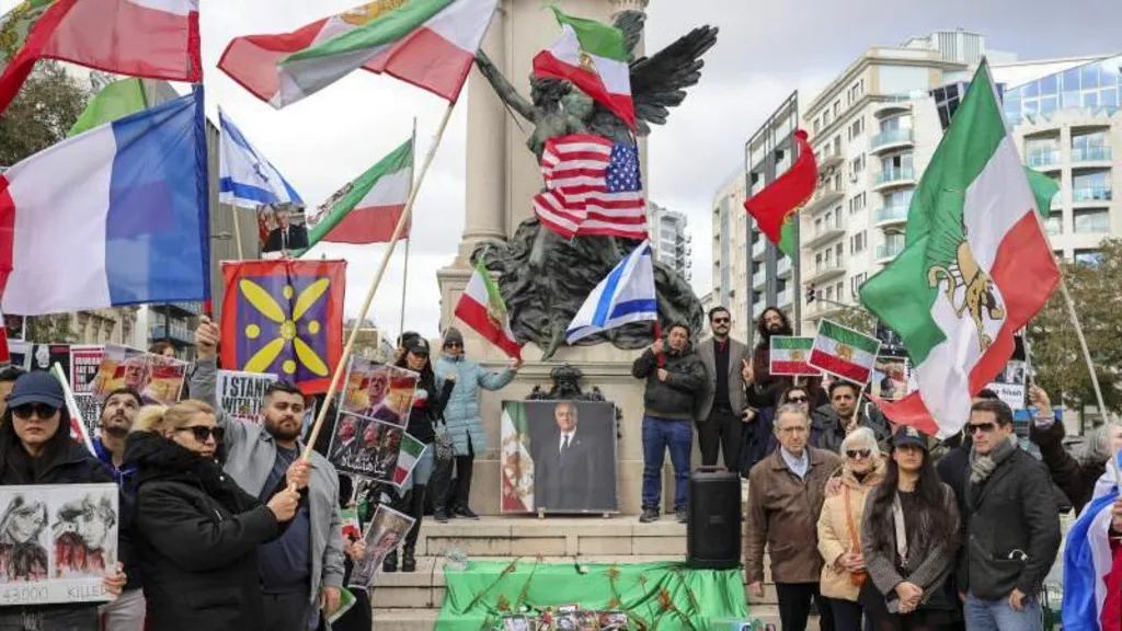 People gather around a picture of Reza Pahlavi. They are holding a variety of signs and flags belonging to several countries including Israel and the United States. 