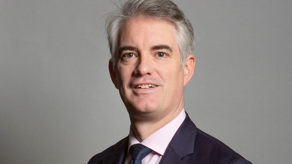 A portrait of Conservative MP for South Suffolk James Cartlidge. He has grey hair and is wearing a navy suit, with a light pink shirt and a navy tie.