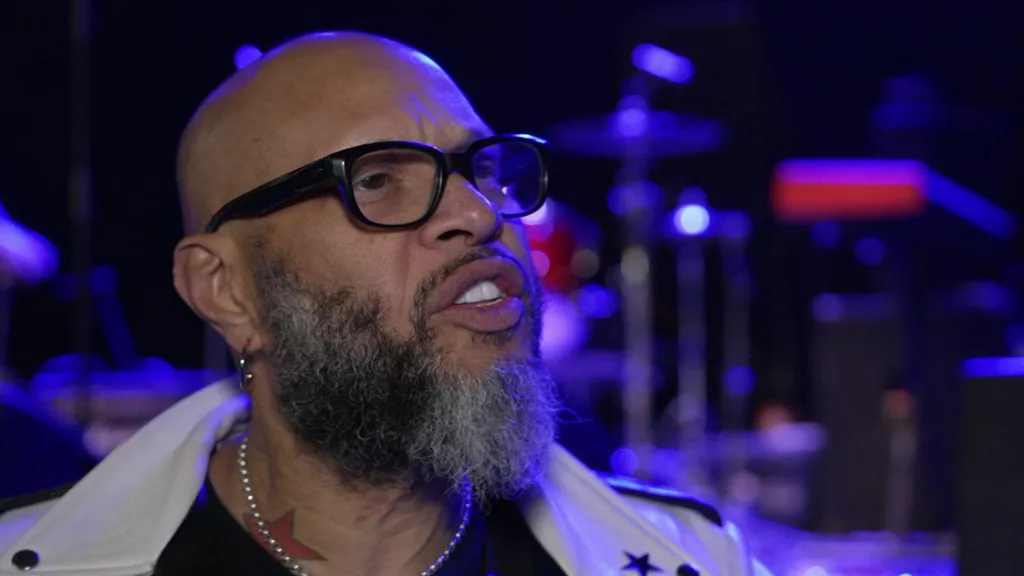 Frank Ferrer gives a TV interview inside a music venue. He is bald with a beard and wears a white leather jacket and jewellery.