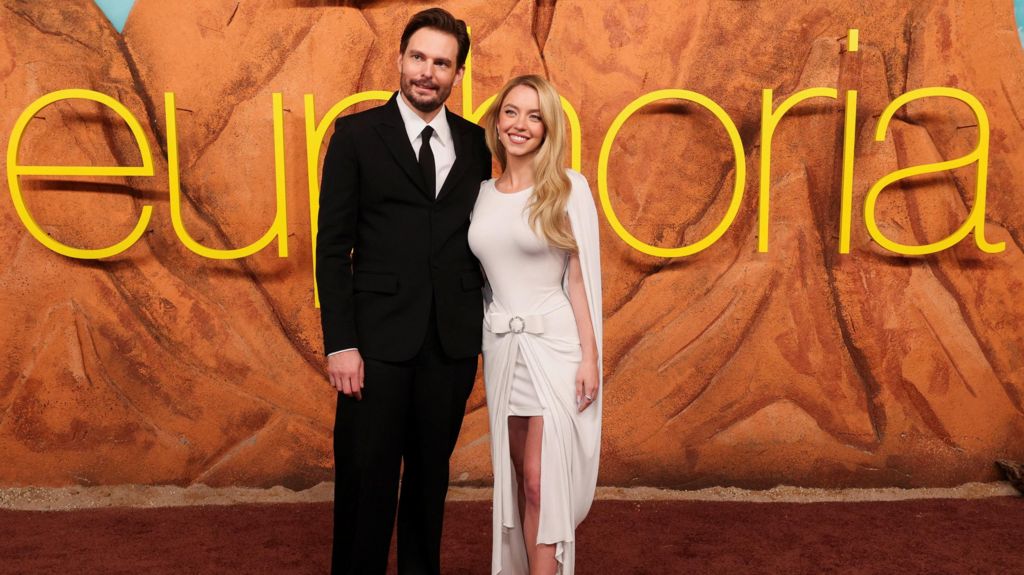 Sydney Sweeney and Sam Levinson standing and smiling together in front of large yellow Euphoria lettering on the red carpet