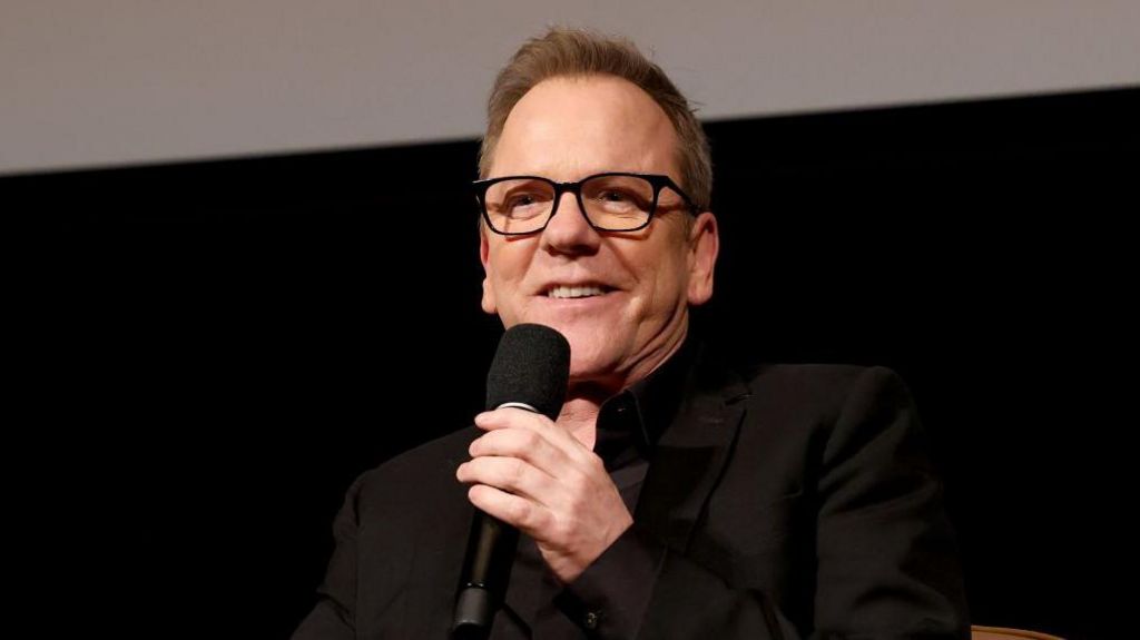 Actor Kiefer Sutherland wearing a black suit and speaking into a microphone
