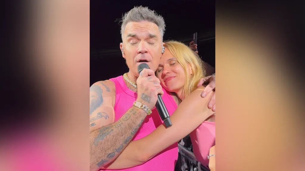 A woman with blonde hair cuddles up to Robbie Williams and has her eyes closed in glee with a smile on her face. Williams wears a pink vest and has his eyes closed as he sings into a microphone