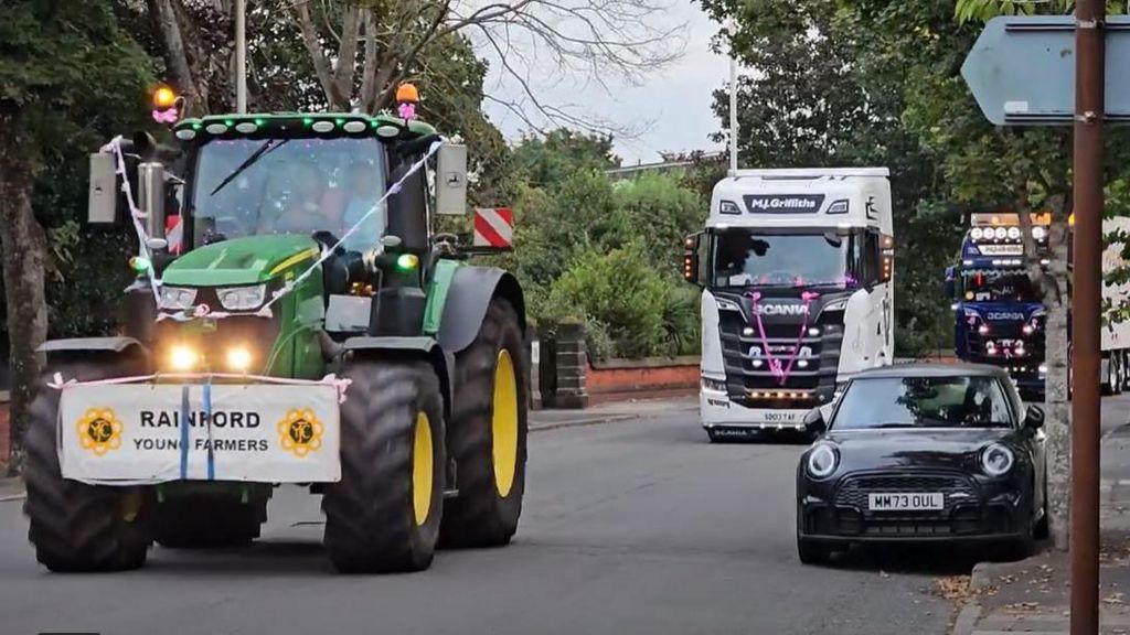 Convoy of tractor and lorries decorated in pink and white ribbons drive down a road