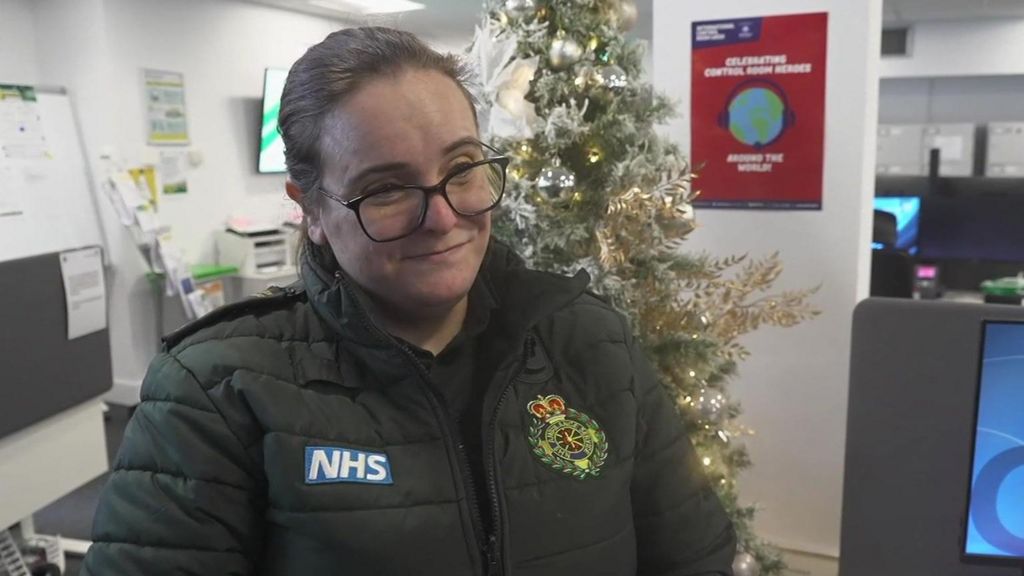An London ambulance worker standing in a call centre. She is wearing a green jacket and there is a Christmas tree behind her. 
