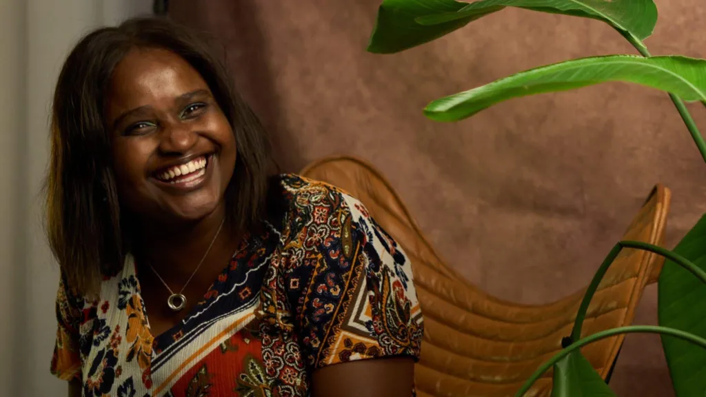 Louise Ndibwirende is smiling, she has shoulder length dark brown hair and wears a patterned top. There is a plant in the foreground, to one side, and she is sat on a brown leather chair with a brown fabric sheet behind her. 
