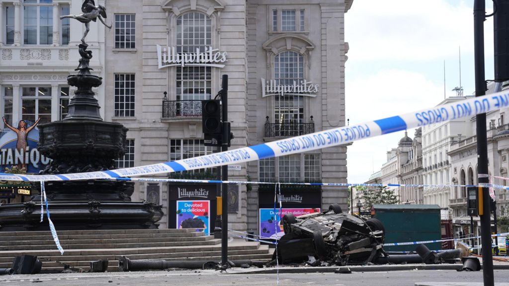A badly damaged black car on its roof in front of a shop building with a sign reading "Lillywhites".