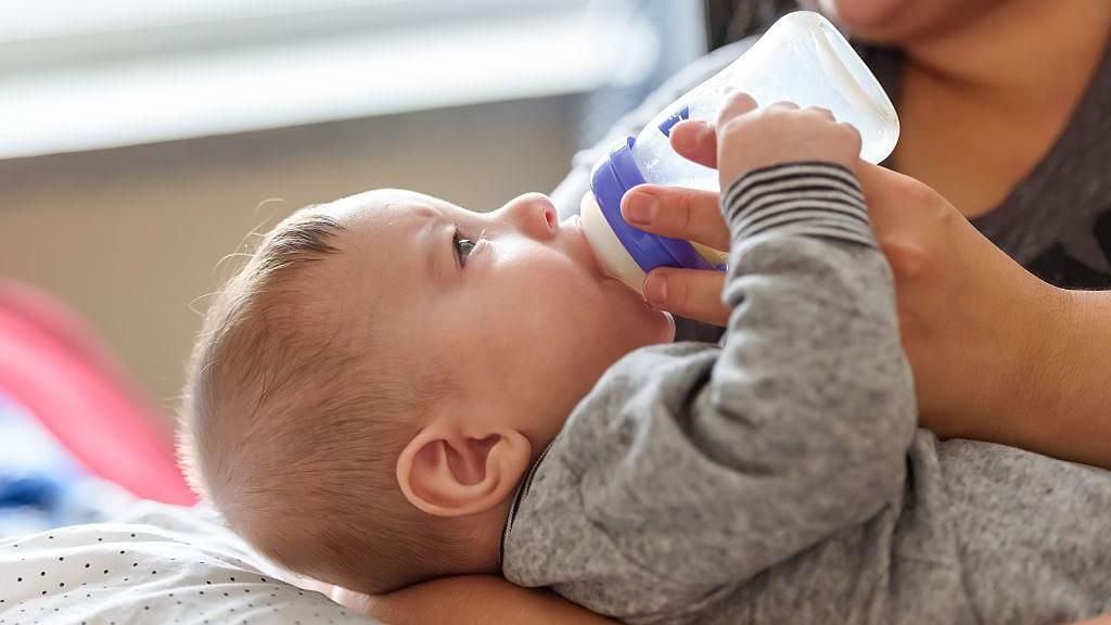 A baby dressed in a grey sleepsuit being held in a woman's lap. The baby is holding the woman's index finger while drinking from a bottle.