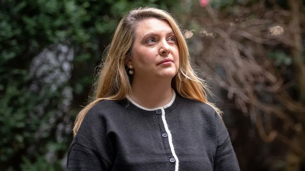 Sophie has long blonde and stands outside in soft natural light, wearing a dark gray buttoned sweater with white trim. Green foliage appears on one side of the background and bare branches on the other.
