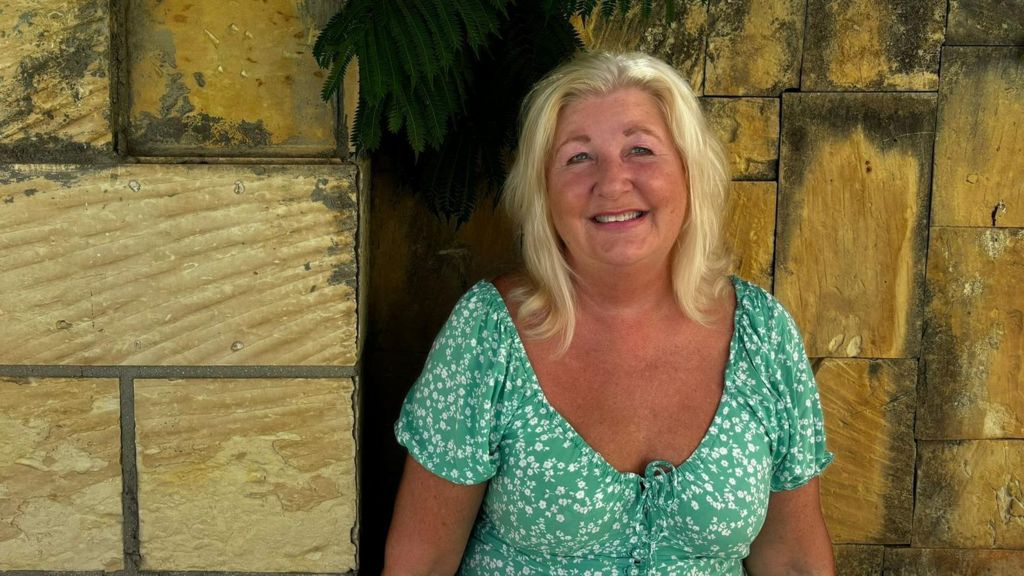 Nicky Maidment is wearing a green milkmaid-style dress with white flowers on it. She has shoulder-length blonde hair and looks tanned, smiling at the camera while perched near a sandy-coloured brick wall with greenery coming out of it.