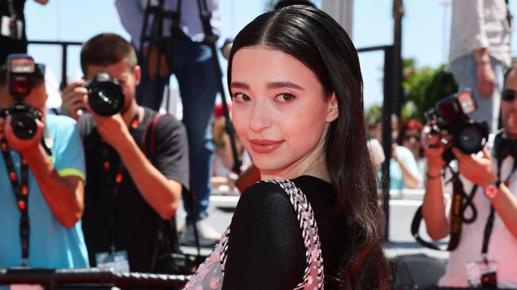 Mikey Madison at the Cannes Film Festival, she is smiling on the red carpet and looking over her shoulder, in a black dress 