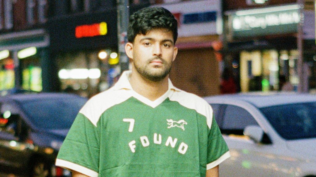 Sufiyaan Salam wearing a green and white T-shirt with the Found logo, standing at dusk in front of cars and restaurants on the Cutty Mile