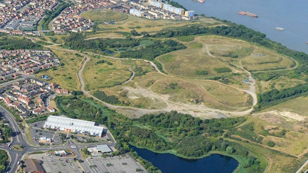 The river Thames can be seen on the right from an aerial shot - with green fields and trees, on the outskirts of which are low and high-rise housing and buildings with a lake in the foreground.
