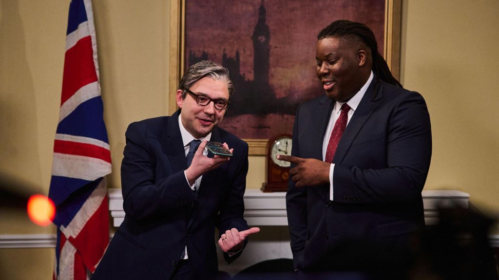 Two actors in a set resembling Ten Downing Street - one is appearing as Keir Starmer, the other as David Lammy, both wearing suits. A Union Flag and painting of Big Ben are behind them
