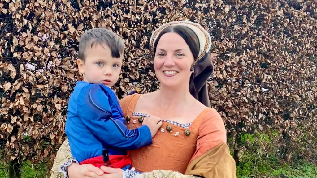 Hollie dressed in a medieval costume as she holds her son Max in her arms. Max is also wearing a costume, with a blue long-sleeved top and red belt.