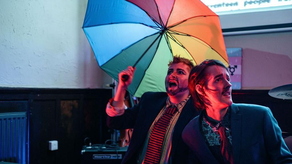 Two drag kings (women dressed as men) with a rainbow umbrella
