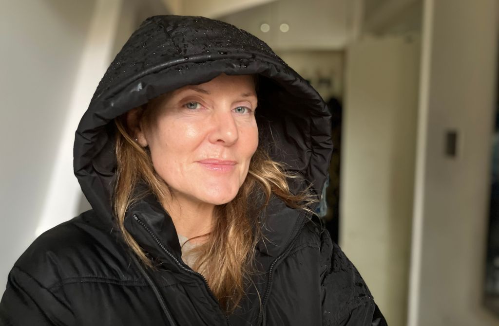 A woman with long, light coloured hair in a black coat with the hood up is interviewed in a car park in the rain. There are cars parked behind her.