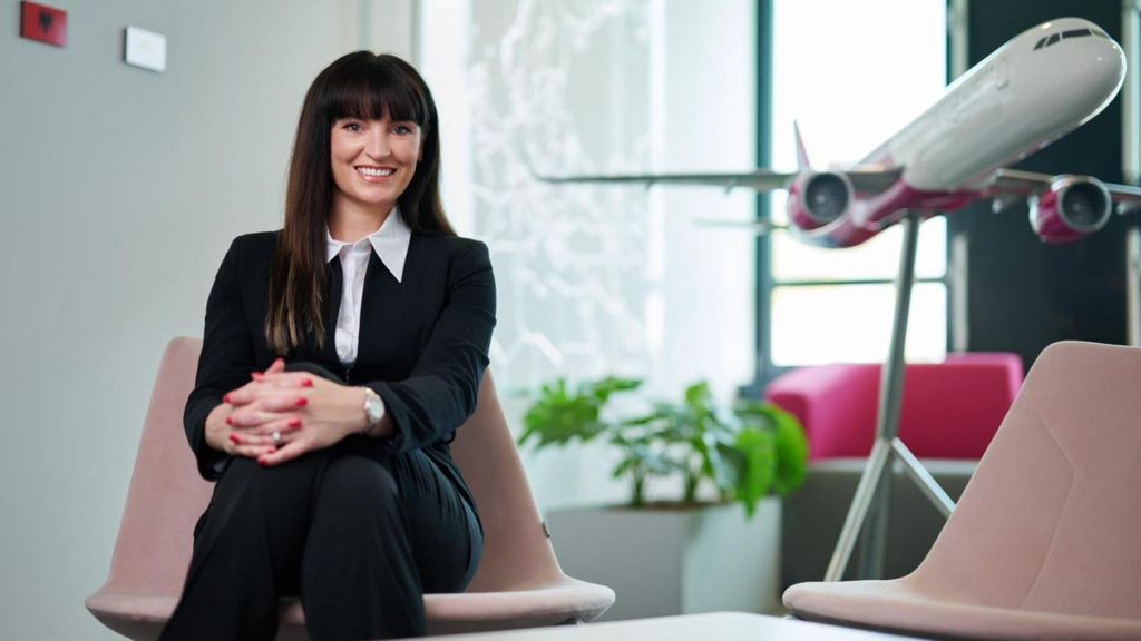 Yvonne smiling while wearing a suit and sitting in a pale pink chair, in front of a large model of an airplane.