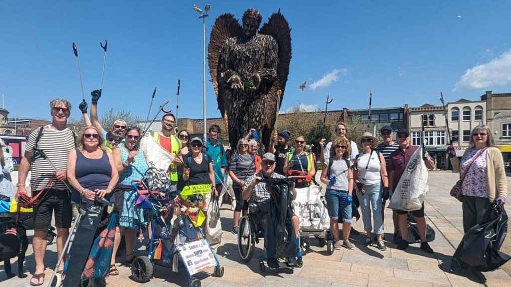 A large group of litter pickers standing in front of the Weston-super-Mare knife angel. Some are wearing hi-vis vests and others are holding bin bags and litter picking tools