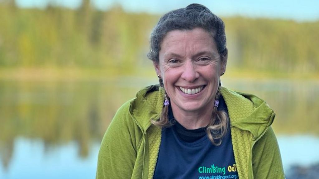 Kelda Wood in a green hoodie and blue tshirt, smiling in front of a lake