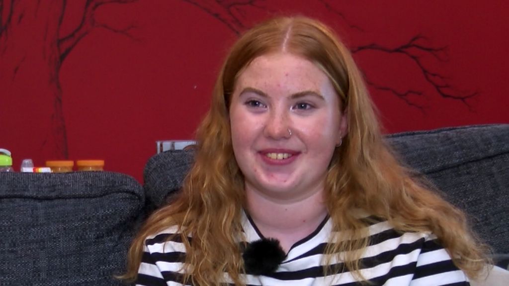 A young girl with ginger hair and a black and white horizontally-striped top smiles while sitting on a couch