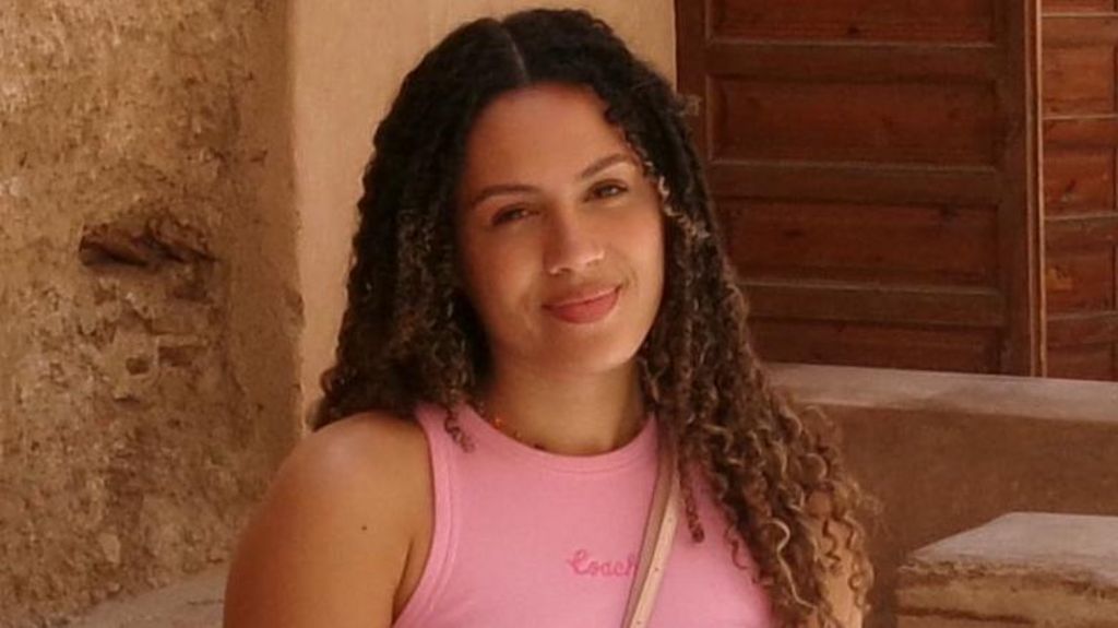 Rochelle, a woman with long curly brown hair in a middle parting, wearing a pink vest top