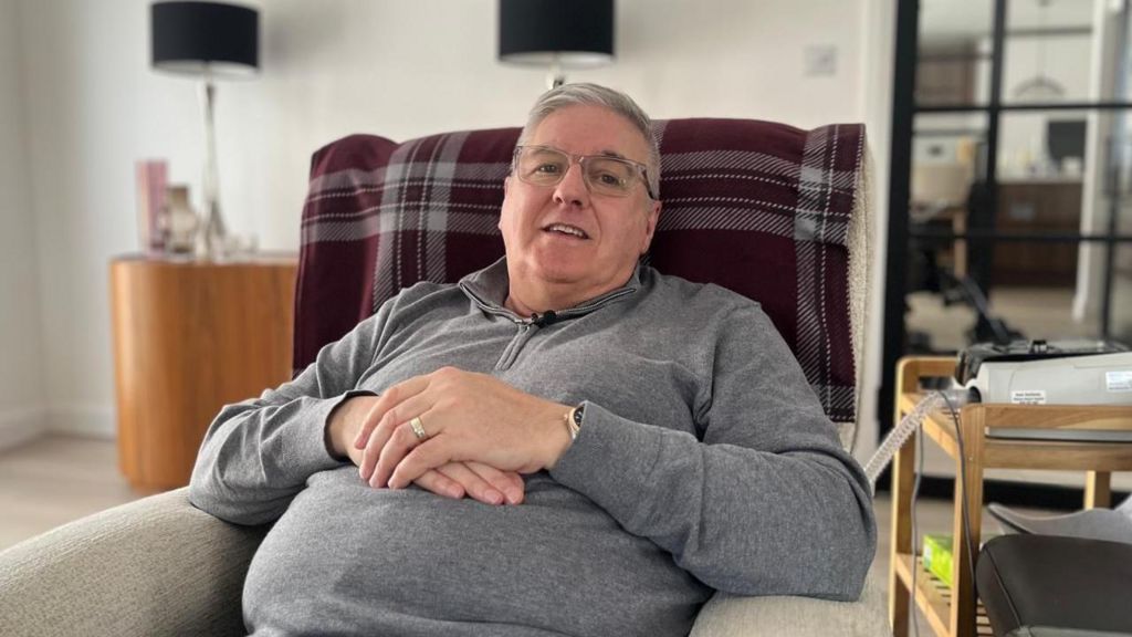 Steven Barrett sits in an armchair in a living room, facing the camera and smiling. He is wearing a grey fleece and glasses.