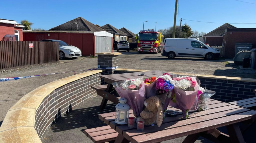 Teddies, flowers and candles have been left on a picnic bench.  Behind, the police tape is attached to a wall and a fire engine is parked.