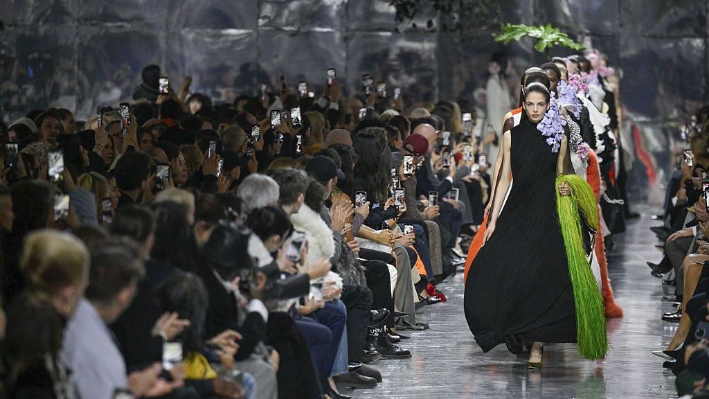 Models walking down a mirrored runway, the first wearing a black draped sleeveless dress and holding a lime green fringed bag - people watching the show are applauding, many of them holding up their phones to take pictures