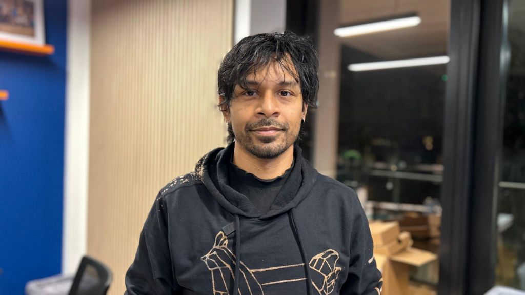 Man wearing a dark grey hoodie looks at the camera. He has black hair, slight brushed forward fringe and a short beard growth. He is in what looks like an office, with a window looking out to a balcony.