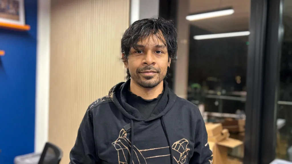 Man wearing a dark grey hoodie looks at the camera. He has black hair, slight brushed forward fringe and a short beard growth. He is in what looks like an office, with a window looking out to a balcony.
