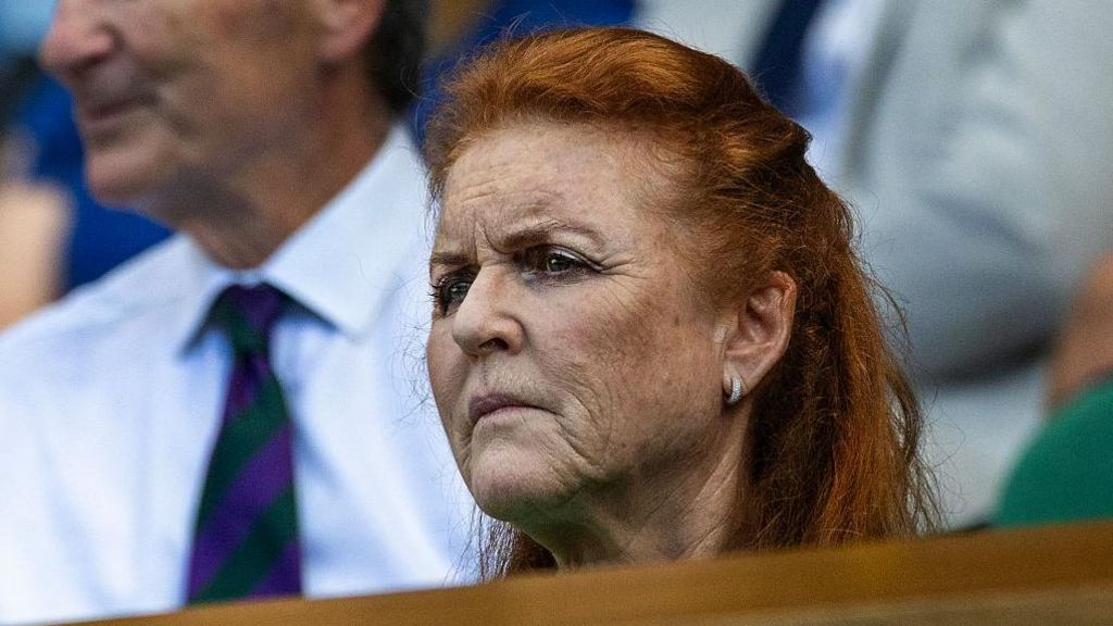 Close up of Sarah Ferguson frowning. In the background there is a man in a white shirt and tie.