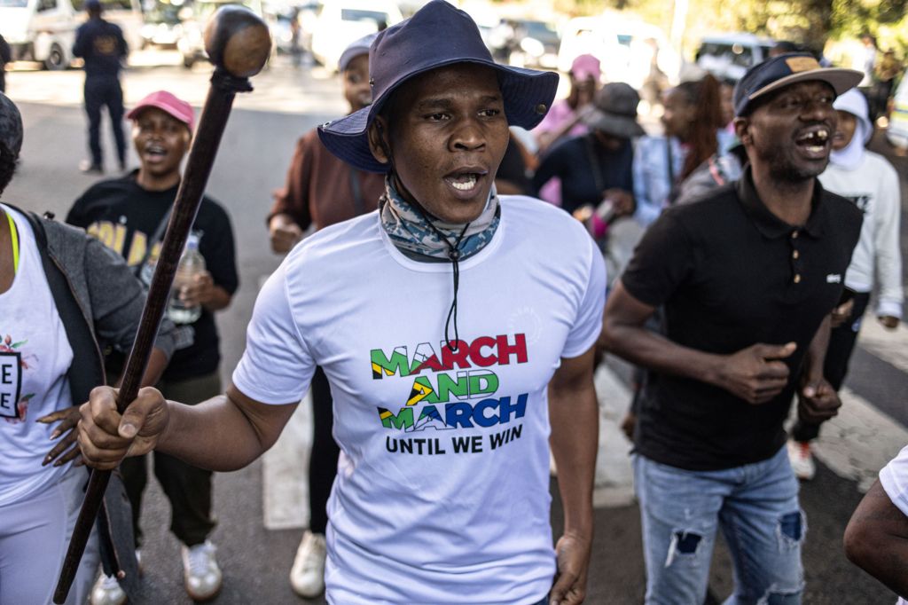 A man wearing a white T-shirt with the slogan: March and March until we win over a South African flag, and holding a stick, on an anti-migrant march